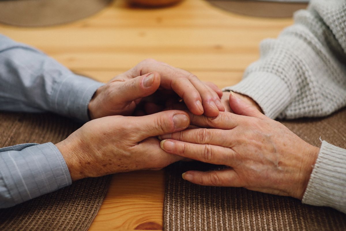 An older person's hands rest folded over a younger person's hands on a wooden table.